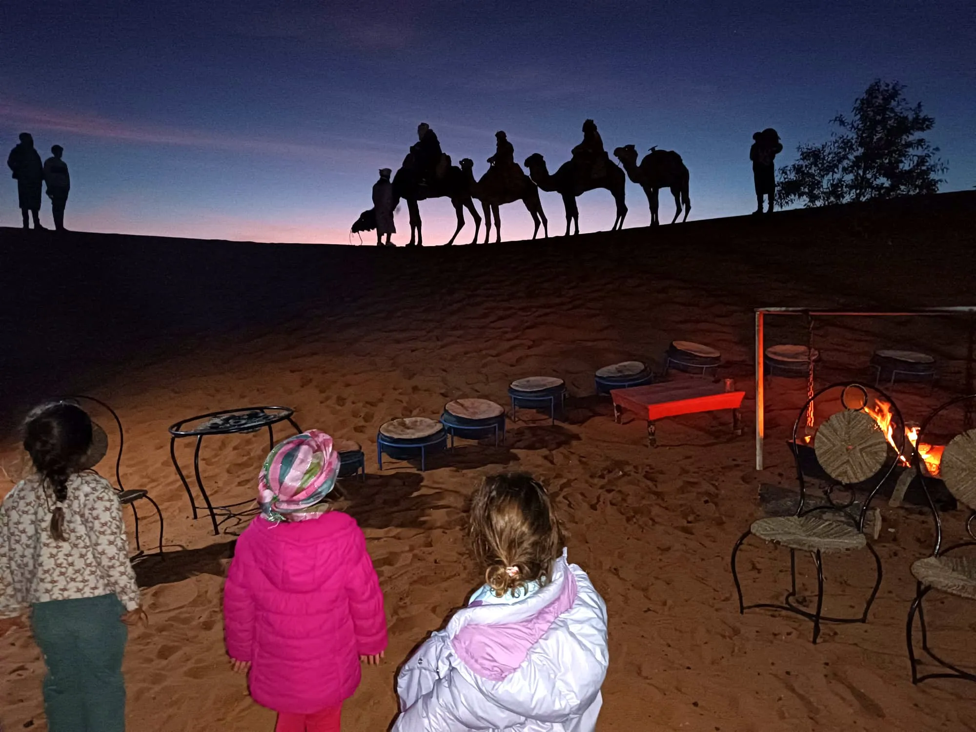 Nenes al costat del foc en un campament del desert al vespre, amb una caravana de dromedaris a l’horitzó